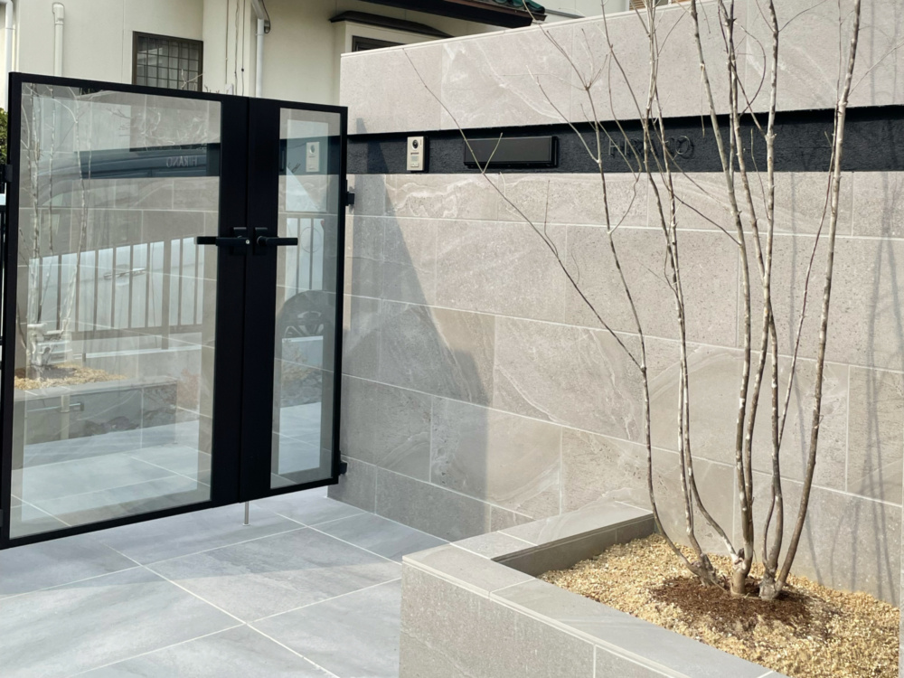愛知県名古屋市緑区にて住友不動産の新築外構工事が完成しました。シャッターゲート付きのクローズ外構とし、門塀を作成し一面を統一して天然石タイル貼り仕上げとし、高級感にある外構エクステリアデザインとなりました。門扉はYKKのルシアス門扉P01型をセレクト。駐車場エリアのカーポートにはYKKのエフルージュをセレクトし設置しています。階段をタイル貼り仕上げの踊り場階段のように配置し、グレーとブラックを基調としたスタイリッシュなモダンスタイルの外構が完成しました。ミライエエクステリア　名古屋市名東区名東店、一宮店